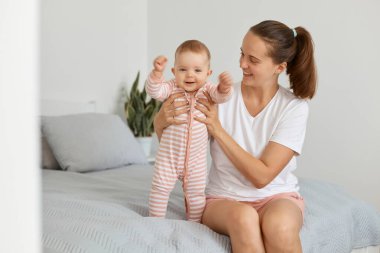 Olumlu iyimser genç annenin portresi, beyaz bir tişört giymiş, bebek bebekle yatakta oturuyor, çocuğun elini tutan kadın, mutlu bir ifadeyle annesinin yanında duran çocuk..