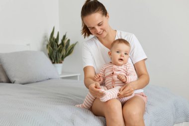 Beyaz atkuyruklu, siyah saçlı, beyaz tişörtlü, beyaz saçlı, bebekle yatakta oturan, annesinin çocuğun ayak parmaklarına dokunan, güzel çocuğuyla oynayan beyaz saçlı yetişkin bir kadın..