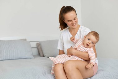 Genç ve mutlu bir annenin beyaz bir tişört giyip bebek bebekle yatakta oturduğu, annesinin sevimli bir gülümsemeyle çocuğa baktığı, çocuğuyla vakit geçirmekten memnun olduğu yatay bir fotoğraf..