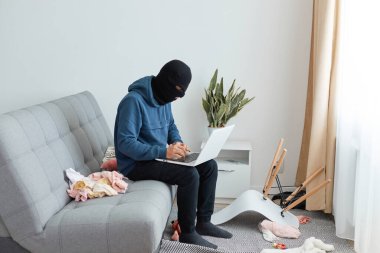 Mavi kapüşonlu ve siyah hırsız maskeli bir adamın portresi, patlak lastik, kanepede oturup dizüstü bilgisayarı açmak, modern tekniği çalmak, defterde önemli dosyalar bulmak,.