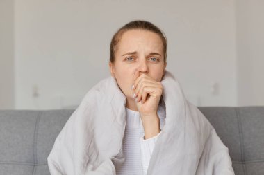 Yorganın altında oturan, öksüren, el ele tutuşan, üzgün yüz ifadesi olan, grip belirtileri gösteren hasta bir kadının portresi..