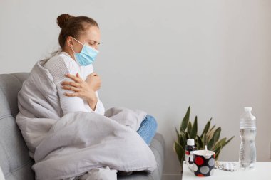 Hasta bir kadının yan görünüşü grip, soğuk algınlığı ya da koronavirüs, kanepeye sarılmış ve tıbbi maske takmış, kendini iyi hissetmiyor, yüksek sıcaklıkta acı çekiyor..