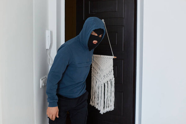 Крытый снимок грабителя в синей толстовке и черной маске, ломающего квартиру, чтобы что-то украсть, заходящего внутрь за кражу чужого дома, опасного ограбления.