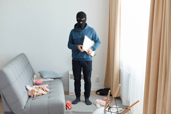 Человек крадет ноутбук в чужом доме, убегает, будучи преследуемым, носит маску грабителя и синюю толстовку, стоит рядом с диваном и окном, опасное ограбление ломается.