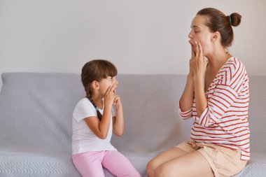Çizgili gömlek ve şort giyen genç beyaz kadın portresi. Kanepede oturan küçük kız, kadın ve çocuk oral kas fonetik hedeflerini yeniden yapılandırmak için egzersiz yapıyorlar..