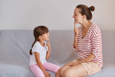 Koyu renk saçlı, çizgili gömlek ve şort giyen bir kadının kanepeye küçük bir kızla oturması konuşma terapisti ve hasta yetiştirme eklemleri, fonetik ders..