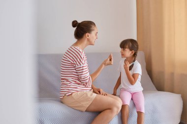Çizgili gömlek ve şort giyen bir kadın, küçük bir kızla koltukta oturuyor, evde derste konuşma egzersizi yapıyor, anne ya da öğretmen ve kadın çocuk birlikte telaffuz etmeyi öğreniyor..