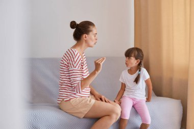 Bun saçlı, çizgili tişörtlü ve şortlu bir kadının kanepede kızıyla oturması, anaokulu öğrencisi bir kızın kadın konuşma terapistiyle doğru telaffuz çalışması..