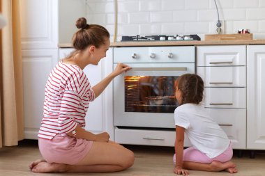 Çizgili tişört giyen bir kadın, siyah saçlı ve örgülü kızı gaz sobasının yanında oturmuş pasta ya da pasta pişiriyor ve birlikte vakit geçiriyorlar..
