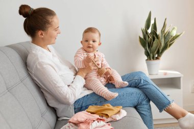 Beyaz tişört ve kot pantolon giyen, bembeyaz saçlı gülümseyen bir kadının bebek kızıyla koltukta oturması, bebeğe bakıp gülümsemesi..