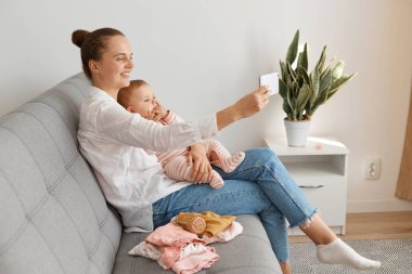 Beyaz gömlek ve kot pantolon giyen, kanepede kızıyla oturan, akıllı telefon tutan ve küçük çocuğuyla selfie çeken bir kadın..