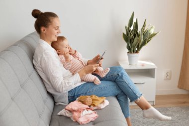 Beyaz gömlek ve kot pantolon giyen, bebek kızıyla koltukta oturan, cep telefonu kullanan, sosyal ağları kontrol eden ya da internette gezinen pozitif kadınların yan görünüm portresi.