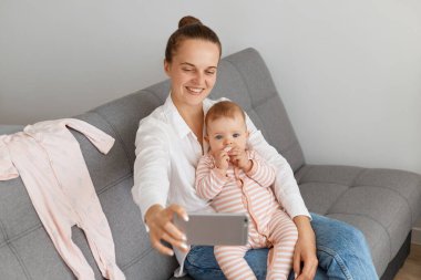 Beyaz gömlekli, kot pantolonlu, gülümseyen beyaz bir kadın. Küçük kızıyla koltukta oturuyor, cep telefonunu tutuyor ve küçük çocuğuyla selfie çekiyor..