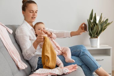 Beyaz gömlek ve kot pantolon giyen bir kadın blogcunun bebek kızıyla kanepede oturup çocuk kıyafetlerini annesinin video bloğundaki takipçilere gösterirken çekilmiş bir fotoğrafı..