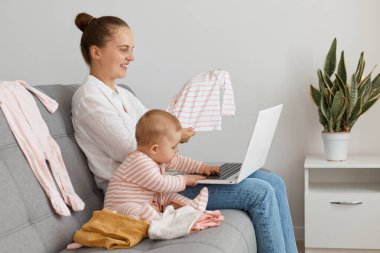 Beyaz gömlek ve kot pantolon giyen çekici bir kadının profil fotoğrafı. Küçük kızıyla kanepeye oturmuş, elinde defter, elinde çocuk kıyafetleri, arkadaşına aldıklarıyla böbürlenirken..