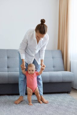 Bun saçlı, beyaz gömlek ve kot giyen, küçük kızına gitmeyi öğreten, arka planda gri öksürükle halıya basan kadın portresi..