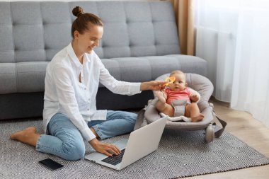 Beyaz gömlek ve kot pantolon giyen, evde oturmuş dizüstü bilgisayarda çalışırken poz veren ve sallanan sandalyedeki çocuğa oyuncağını gösteren iç mekanda gülümseyen mutlu bir kadın..