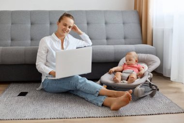 Gevşemiş, iyimser bir kadın. Bun saç stili, beyaz gömlek ve kot giyen, kızının yanında poz veren, elinde not defteri olan, ekrana bakan, film izleyen..