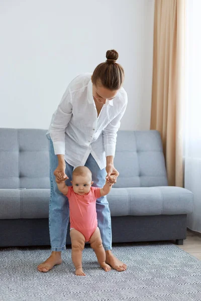 Bun saçlı, beyaz gömlek ve kot giyen, küçük kızına gitmeyi öğreten, arka planda gri öksürükle halıya basan kadın portresi..