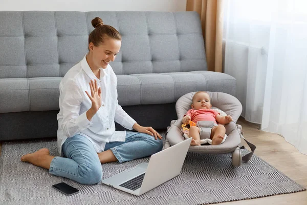 Bun saçlı, beyaz gömlekli, kot pantolonlu, yerde yeni doğmuş bir bebekle oturan ve dizüstü bilgisayarında video görüşmesi yapan, el sallayan, merhaba diyen mutlu bir kadının yatay fotoğrafı..