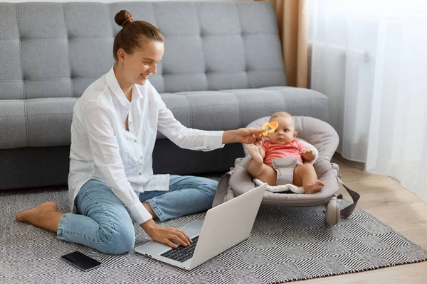 Beyaz gömlek ve kot pantolon giyen, evde oturmuş dizüstü bilgisayarda çalışırken poz veren ve sallanan sandalyedeki çocuğa oyuncağını gösteren iç mekanda gülümseyen mutlu bir kadın..