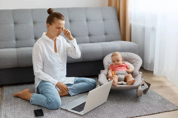 Bun saçlı, beyaz gömlek ve kot giyen, bebekle oynayan ve internetteki işini yapan yorgun bir kadının portresi, gözlerinde acı hisseder, gözünü ovar..