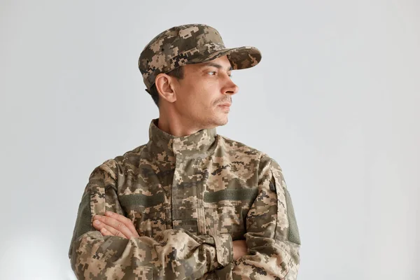 Image of young adult serious military man wearing camouflage uniform and hat, standing with folded hands, having confident facial expression, looking away.