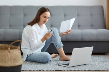 Beyaz gömlek ve kot pantolon giyen, elinde kağıt belge ve cep telefonu olan ve faturaları internet üzerinden ödeyen genç bir kadının resmi..