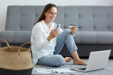 Beyaz gömlekli, kot pantolonlu, elinde akıllı telefon ve kredi kartıyla yerde oturan mutlu beyaz kadın, olumlu duygularını dile getiriyor, internetten ödeme yapıyor..