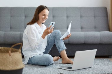 Beyaz gömlek ve kot pantolon giyen, elinde kağıt ve akıllı telefonuyla yerde oturan, internet üzerinden çalışan ya da internet üzerinden fatura ödeyen mutlu bir kadının kapalı çekimleri..
