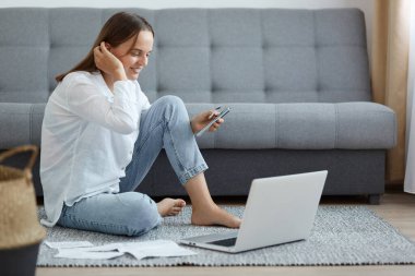 Beyaz gömlek ve kot pantolon giymiş gülümseyen güzel bir kadının yan görünüm portresi, yere oturmuş, pozitif yüz ifadesiyle ekrana bakıyor, saçına dokunuyor, internetten para ödüyor..