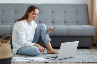 Kafası karışmış, beyaz tişörtlü, kot pantolonlu, cep telefonu ve kredi kartıyla yerde oturan, kafası karışmış, kafası karışmış, kafası karışık, kafası karışık bir kadın resmi..