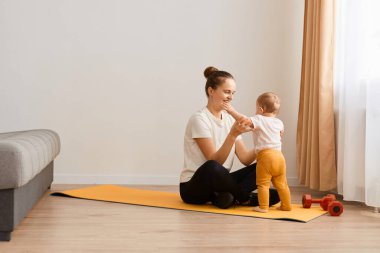Beyaz tişört giyen genç kadın portresi ve siyah tozluklar evde spor yapıyor ve küçük kızıyla oynuyor, kanepe ve pencerenin yanında mindere oturuyor..