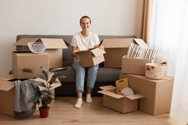 Beyaz t-shirt ve kot pantolon giyen, taşınma sırasında koltukta oturan, karton kutuyla eşyalarını taşıyan, kameraya bakan bir kadının kapalı çekimleri..