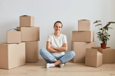 Beyaz t-shirt giyen, etrafı karton kutularla çevrili, mutlu bir ifadeyle kameraya bakan, kollarını kavuşturmuş, gülümseyen olumlu bir kadın..