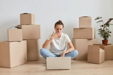 Beyaz tişörtlü bir kadının, etrafı karton kutularla çevrili bir şekilde yerde otururken, laptopta çalışırken, ekrana bakarken, ağzını kapatırken, hata yaparken..