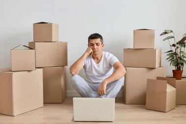 Üzgün, yorgun, beyaz tişörtlü esmer adam etrafı karton kutularla çevrili yerde oturuyor ve dizüstü bilgisayarda çalışıyor, sıkılmış bir ifadeyle görüntüye bakıyor..