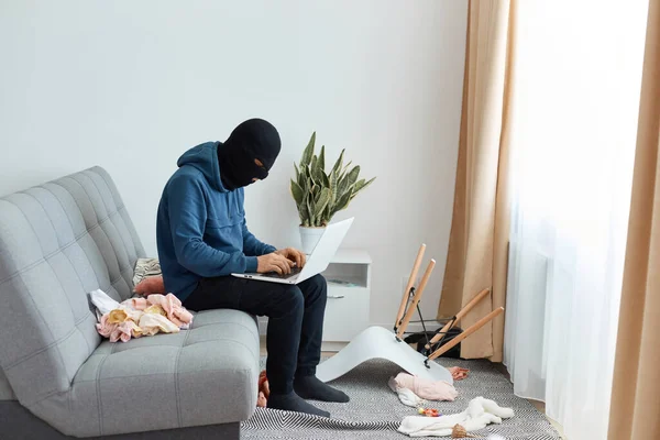 在室内拍摄的身份不明的窃贼身穿蓝色的帽衫 戴着黑色的面具 坐在沙发上 在别人的公寓里用偷来的笔记本电脑 发现了秘密信息 — 图库照片