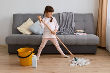 Uzun boylu, siyah saçlı, günlük kıyafetler giyen, kanepenin yanında duran, yerleri yıkayan küçük bir kızın portresi annesinden ev için yardım almak zorunda..