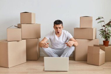 Şok olmuş beyaz tişörtlü, etrafı karton kutularla çevrili ve dizüstü bilgisayarla çalışan korkmuş bir adam hayret verici haberler görüyor..