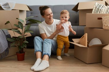 Beyaz tişörtlü bir kadının portresi. Etrafı karton kutularla çevrili ve bebeğiyle oynuyor. Mutluluğu ifade ediyor..