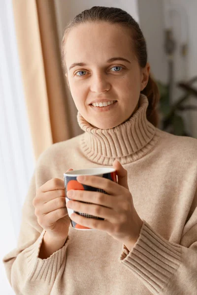 照片中 一位面带微笑的积极女性身穿米黄色毛衣 站在屋里 端着一杯咖啡和茶 面带微笑地看着相机 — 图库照片