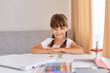 Ev ödevi yapıp kitap okuyan mutlu çocuk portresi, siyah saçlı ve örgülü şirin bir kız çocuğu kameraya pozitif bir ifadeyle bakıyor, uzaktan öğreniyor..