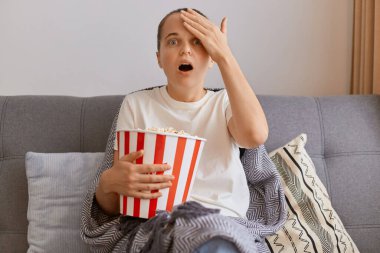 Beyaz tişörtlü, patlamış mısır ve uzaktan kumandayla koltukta oturan, korku filmi seyreden ağzı açık ve çok etkilenmiş genç bir kadının kapalı çekimleri..