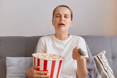 Beyaz t-shirt giyen, elinde patlamış mısır ve uzaktan kumandayla koltukta oturan, romantik film izleyen ya da dizide trajik bir an gören üzgün genç bir kadının kapalı çekimleri..