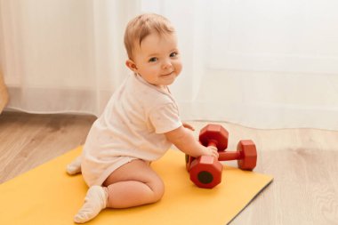 Yoga minderinde oturan, elinde dambıllarla kameraya gülümseyen, spor ekipmanlarını keşfeden sevimli küçük bir çocuğun yatay çekimi..