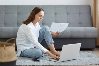 Koyu renk saçlı, beyaz gömlekli, kot pantolonlu, kanepenin yanında oturan, dizüstü bilgisayar kullanan, online proje yapan, elinde kağıt tutan sevimli bir kadın portresi..