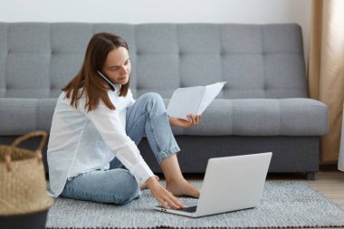 Koyu renk saçlı, beyaz gömlekli ve kot pantolonlu genç bir kadın. Kanepenin yanında oturuyor. Laptop kullanıyor, telefonla konuşuyor, kağıt tutuyor, faturalarla ilgili sorunları çözüyordu..