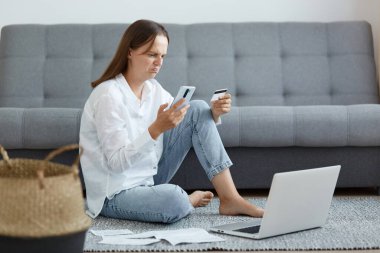 Hüzünlü kadın portresi, beyaz gömlek ve kot pantolon giyen, kanepenin yanında oturan, dizüstü bilgisayar ve telefon kullanan, kredi kartı tutan, veri girerken sorun yaşayan..