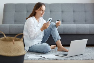 Beyaz gömlek ve kot pantolon giyen, diz üstü bilgisayar ve telefon kullanarak elinde kredi kartı olan beyaz bir kadının yatay görüntüsü..